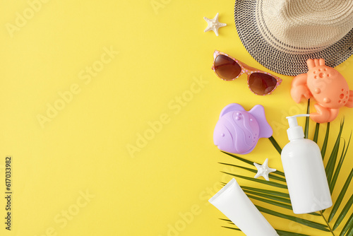 The concept of summer skincare for children. Top view of sunscreen bottles, sun hat, sunglasses, beach toys, palm leaf, starfish on yellow background with blank space for ads or text