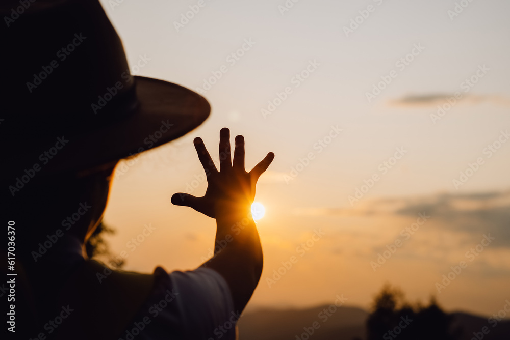 Little girl in hat raising hand over sunset sky, enjoying life and nature. Child on summer field enjoying sunlight. Silhouette of preteen kid in sunlight rays. Environment concept. Dream of flying