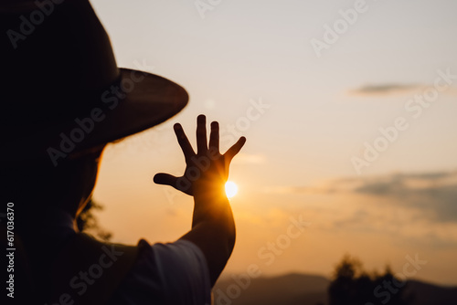 Little girl in hat raising hand over sunset sky, enjoying life and nature. Child on summer field enjoying sunlight. Silhouette of preteen kid in sunlight rays. Environment concept. Dream of flying
