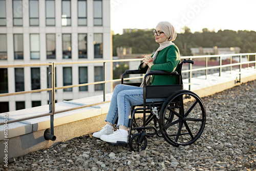 Charming muslim female in traditional headscarf spending morning hours on panoramic terrace in city. Relaxed wheelchair user in casual clothes and eyewear overlooking urban scenery on flat roof. © sofiko14