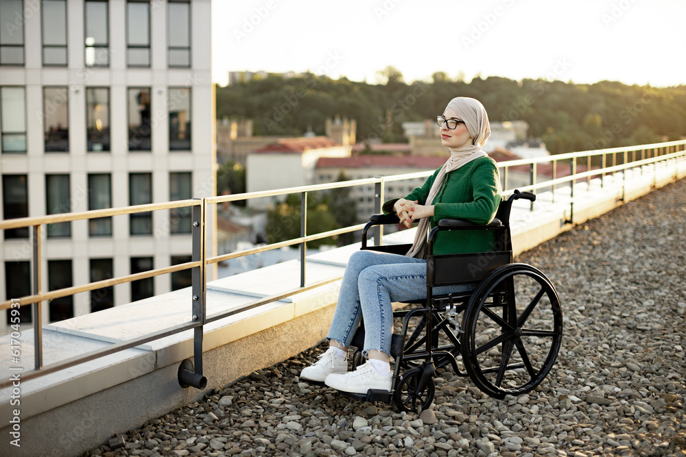 Naklejka premium Full length view of pretty arabian lady in hijab posing in wheelchair on rooftop with cityscape on background. Delighted young woman with mobility impairment promoting healthy outdoor lifestyle.