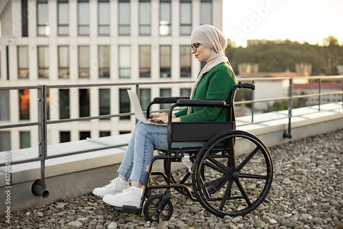 Confident muslim woman in headscarf and glasses typing on laptop while holding modern device on knees on roof terrace. Efficient freelancer in wheelchair doing virtual job in outdoor workplace.