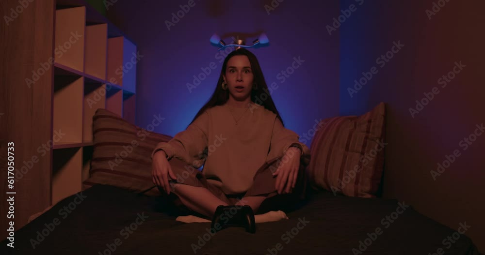 Shocked female gamer playing online game on console with surprised face in blue, looking surprised at camera and controlling game process on gamepad. Shocked female gamer playing online game on console with surprised face in blue, looking surprised at camera and controlling game process on gamepad.