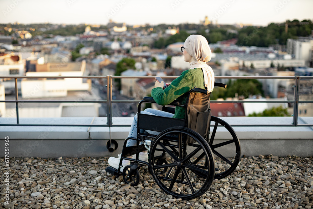 Naklejka premium Charming muslim lady in traditional clothing with cell phone in hands while unwinding on roof terrace. Calm bespectacled woman in wheelchair relaxing while taking in bird's-eye view of city.