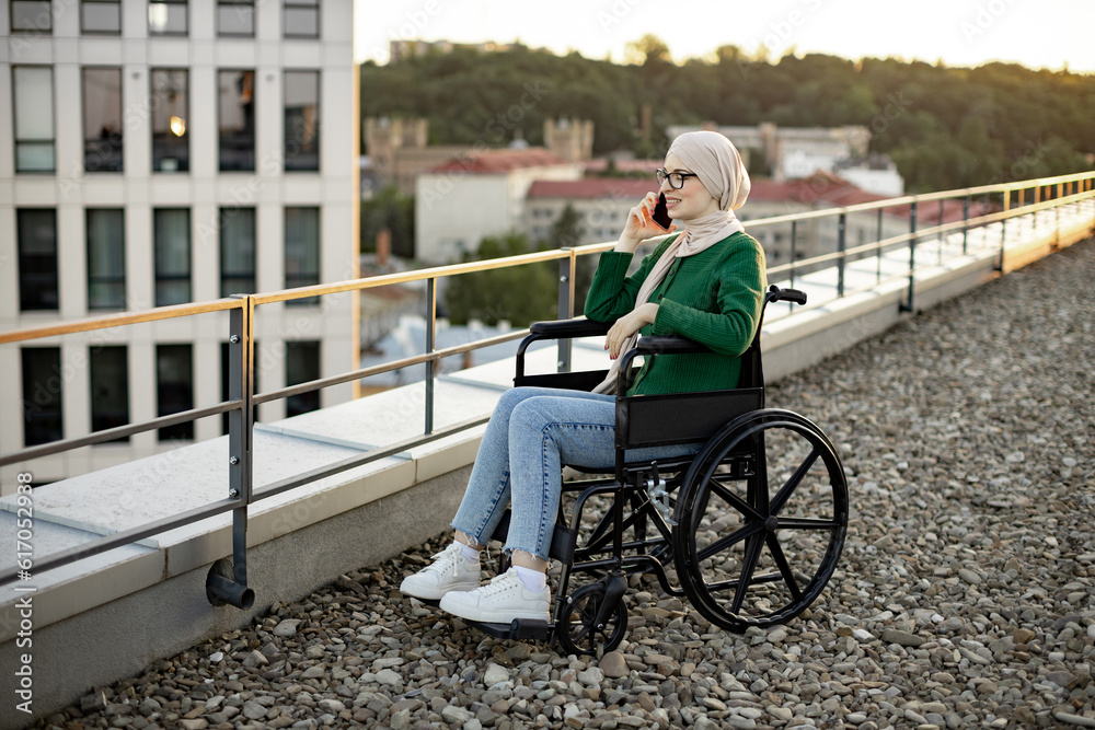 Easygoing muslim person in headscarf chatting on smartphone while sitting in wheelchair on rooftop of city building. Pretty bespectacled lady having friendly conversation with relatives outside home. Easygoing muslim person in headscarf chatting on smartphone while sitting in wheelchair on rooftop of city building. Pretty bespectacled lady having friendly conversation with relatives outside home.