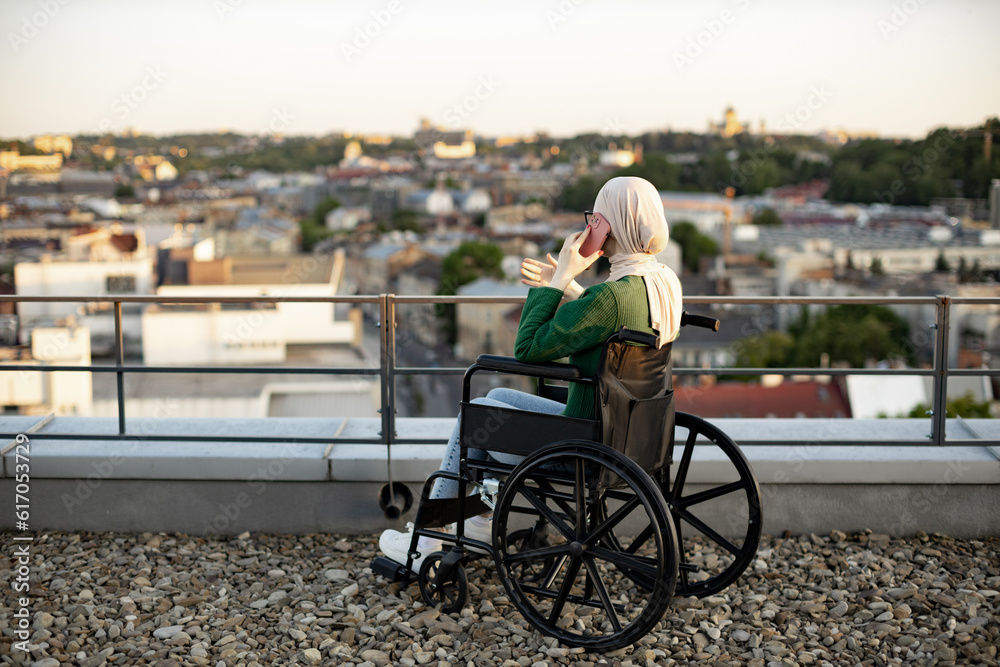 Back view of muslim female in headscarf getting phone call while letting eyes roving over skyline on roof terrace. Relaxed wheelchair user enjoying communication via modern gadget in open air. Back view of muslim female in headscarf getting phone call while letting eyes roving over skyline on roof terrace. Relaxed wheelchair user enjoying communication via modern gadget in open air.