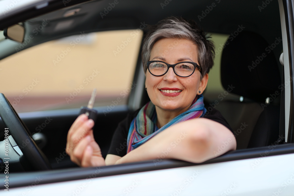 A pretty adult woman in glasses sits at the wheel of a rented car with a key in her hand.