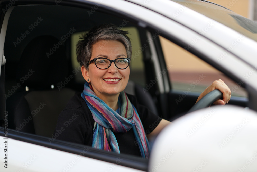 Portrait of a happy adult woman with a short haircut in glasses driving a white car. The concept of renting or buying a car.