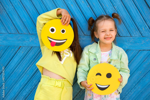 Humorous funny girls hold various funny smiley faces and make faces standing at the blue wall