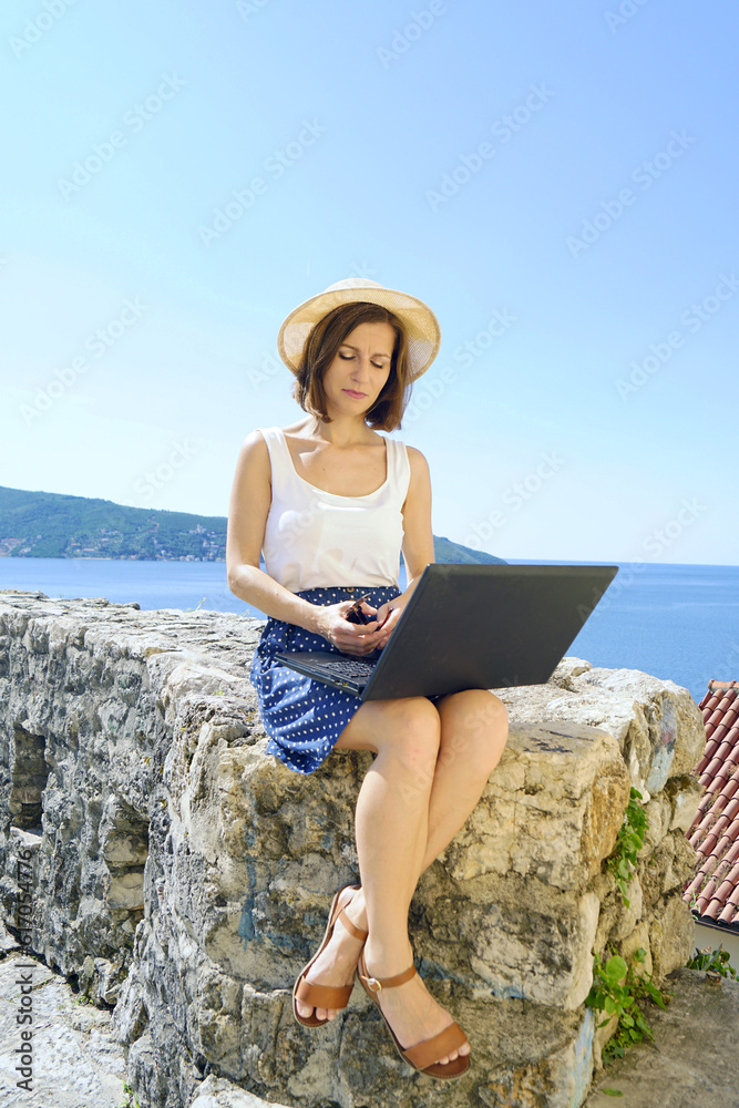 Pretty woman in a hat is working on a laptop, sitting on the background of the sea. Portrait of a freelancer doing remote work while traveling.