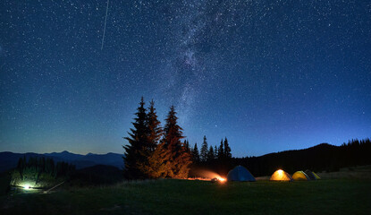 Night camping in mountains under starry sky. Tourist tents in campsite near burning campfire under beautiful sky full of stars with Milky way above forest. Concept of tourism and traveling.