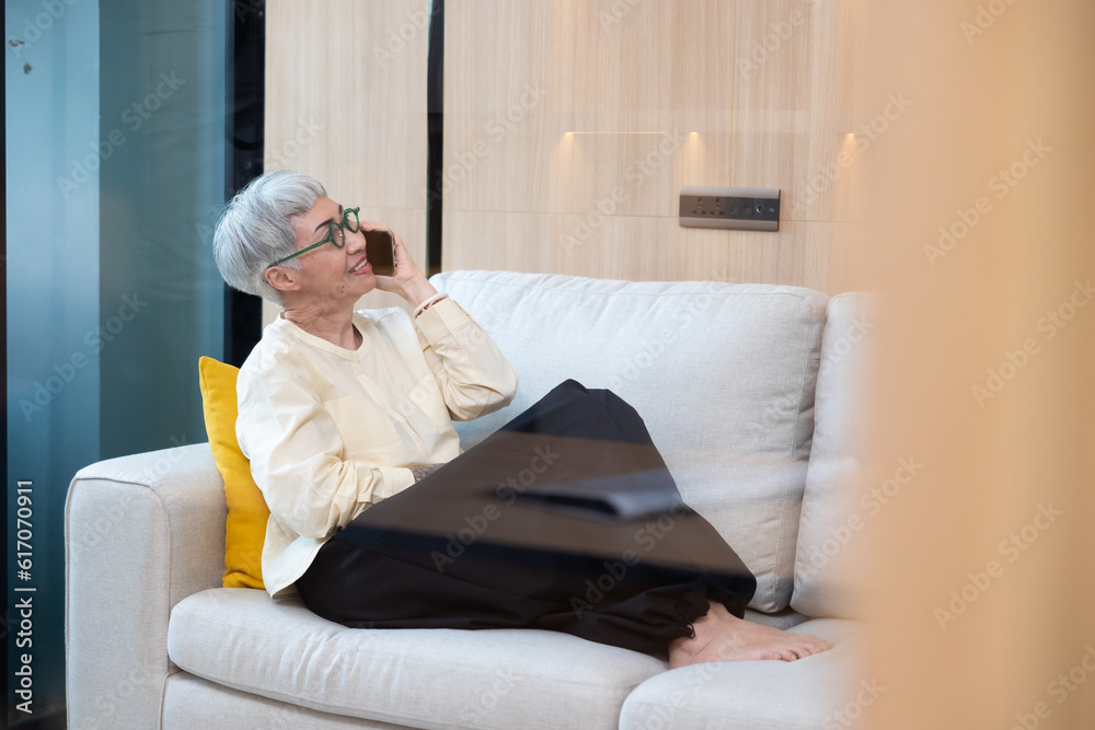 Happy Asian senior mature female holding smartphone using mobile phone technology device sitting on couch at modern home