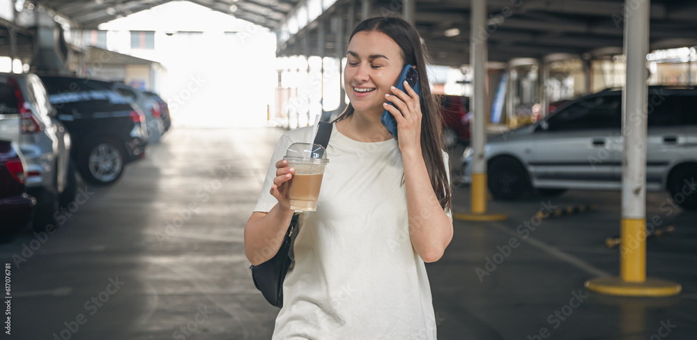 Fototapeta premium A young woman with coffee talking on the phone in the parking lot.