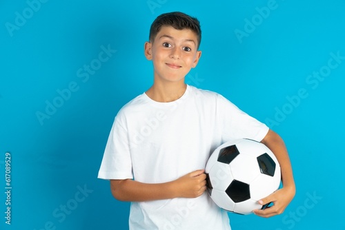 Dreamy rest relaxed Little hispanic boy wearing white T-shirt holding a football ball crossing arms, © Jihan