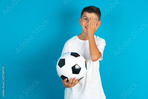 Little hispanic boy wearing white T-shirt holding a football ball covering one eye with her hand, confident smile on face and surprise emotion. © Jihan