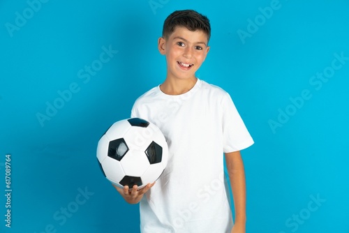 Little hispanic boy wearing white T-shirt holding a football ball winking looking at the camera with sexy expression, cheerful and happy face.