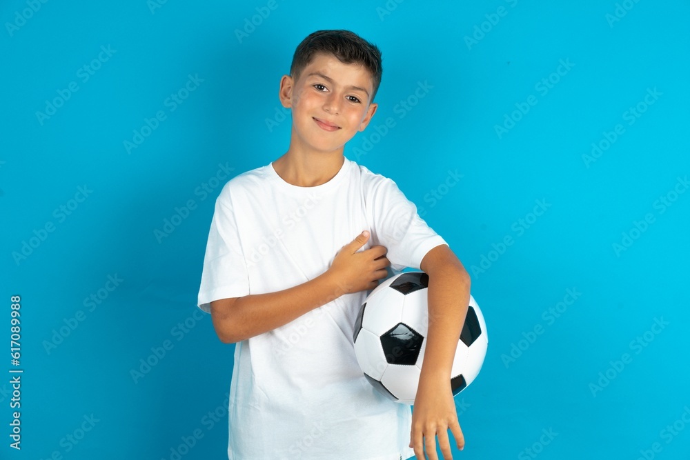 Little hispanic boy wearing white T-shirt holding a football ball happy face smiling with crossed arms looking at the camera. Positive person. Little hispanic boy wearing white T-shirt holding a football ball happy face smiling with crossed arms looking at the camera. Positive person.