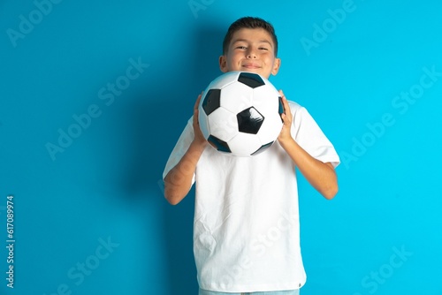 Little hispanic boy wearing white T-shirt holding a football ball praying with hands together asking for forgiveness smiling confident. © Jihan