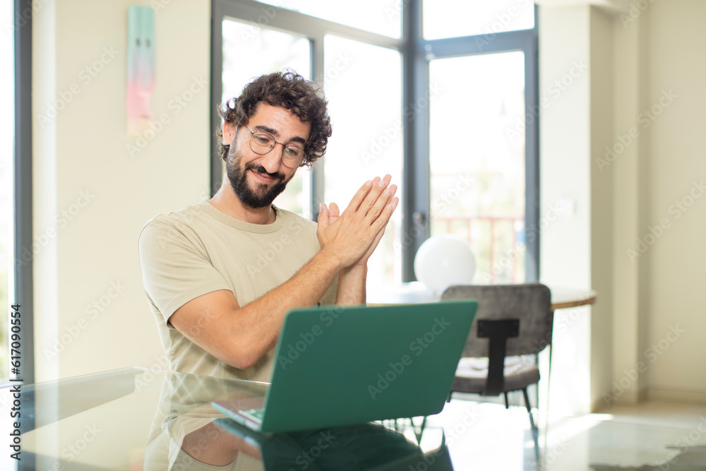 young adult bearded man with a laptop feeling happy and successful, smiling and clapping hands, saying congratulations with an applause young adult bearded man with a laptop feeling happy and successful, smiling and clapping hands, saying congratulations with an applause