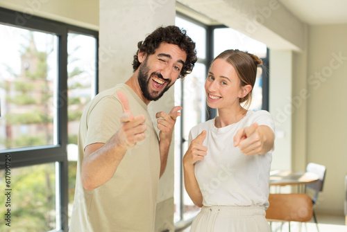 young adult couple feeling happy, cool, satisfied, relaxed and successful, pointing at camera, choosing you