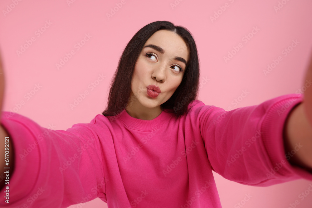 Young woman taking selfie and blowing kiss on pink background Young woman taking selfie and blowing kiss on pink background