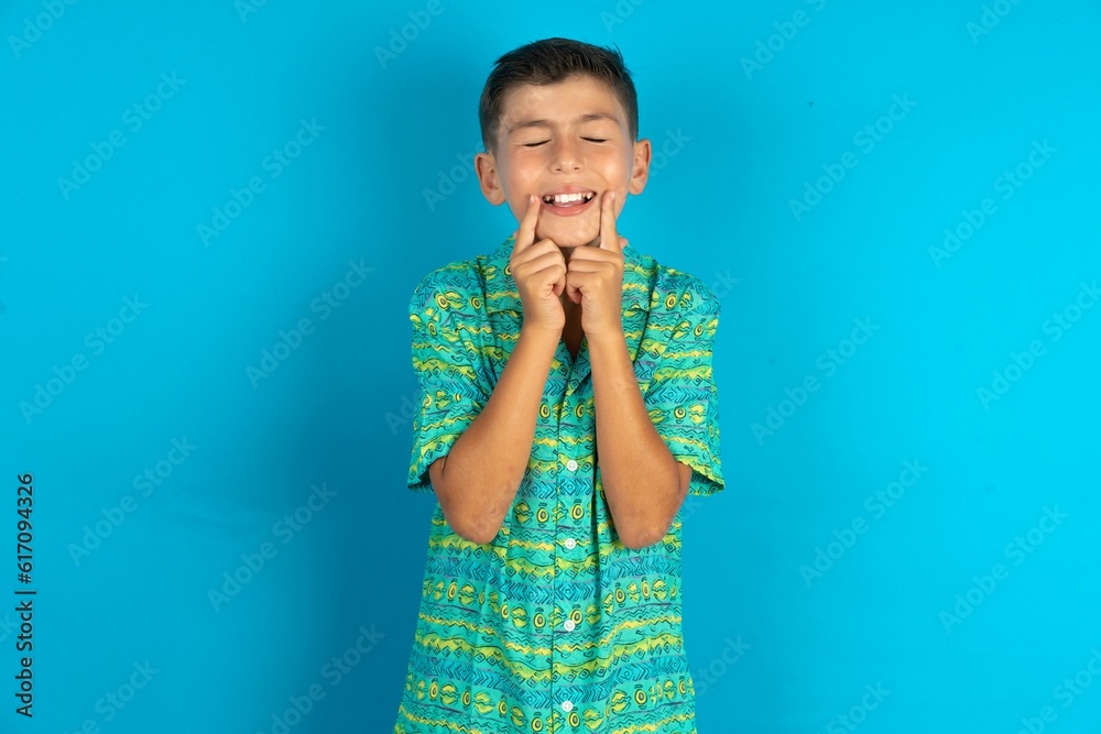Happy Little hispanic boy wearing green aztec shirt with toothy smile, keeps index fingers near mouth, fingers pointing and forcing cheerful smile Happy Little hispanic boy wearing green aztec shirt with toothy smile, keeps index fingers near mouth, fingers pointing and forcing cheerful smile