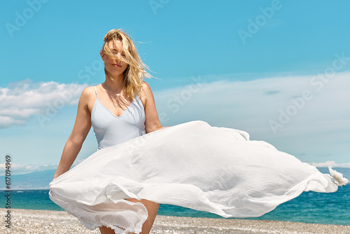 Pretty blond woman in swimsuit and white pareo enjoying sunny windy day near blue sea. Happy female enjoying the life. Beach summer  travel concept.