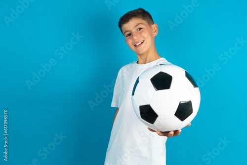 Little hispanic boy wearing white T-shirt holding a soccer ball smiling cheerful offering palm hand giving assistance and acceptance.