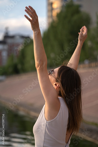 Urban summer portrait of young brunette woman. A woman stands on the river bank with her hands up.