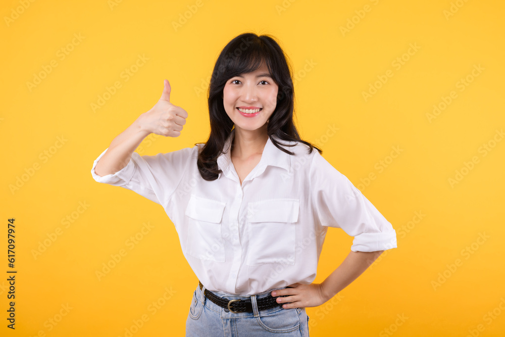 Young asian business woman, smiling while showing thumbs up gesture, recommending product, praise, standing over yellow studio background. Businesswoman confident hand gesture concept. Young asian business woman, smiling while showing thumbs up gesture, recommending product, praise, standing over yellow studio background. Businesswoman confident hand gesture concept.