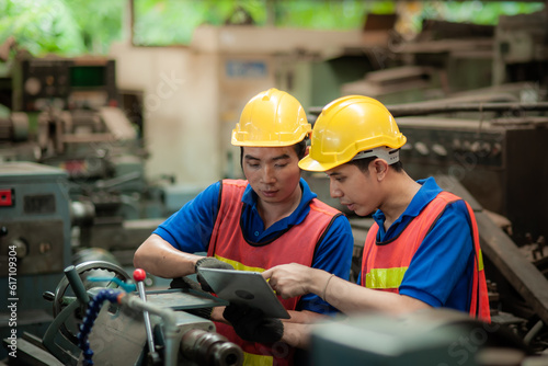 Two young asian male technicians wearing safety uniform workwear, helmets, vest and gloves are Working on a metal lathe and inspecting parts intently In industrial plants.