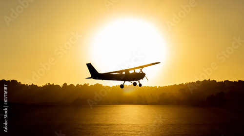 Backlit silhouette of a cessna plane outlined with the sun just behind it flying low with the sea water below illuminated by the golden light of sunset and trees and vegetation in the background © sirbouman