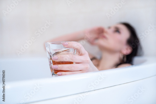 Class of strong alcohol in female hand that belongs young brunette, with makeup lying in bathroom.