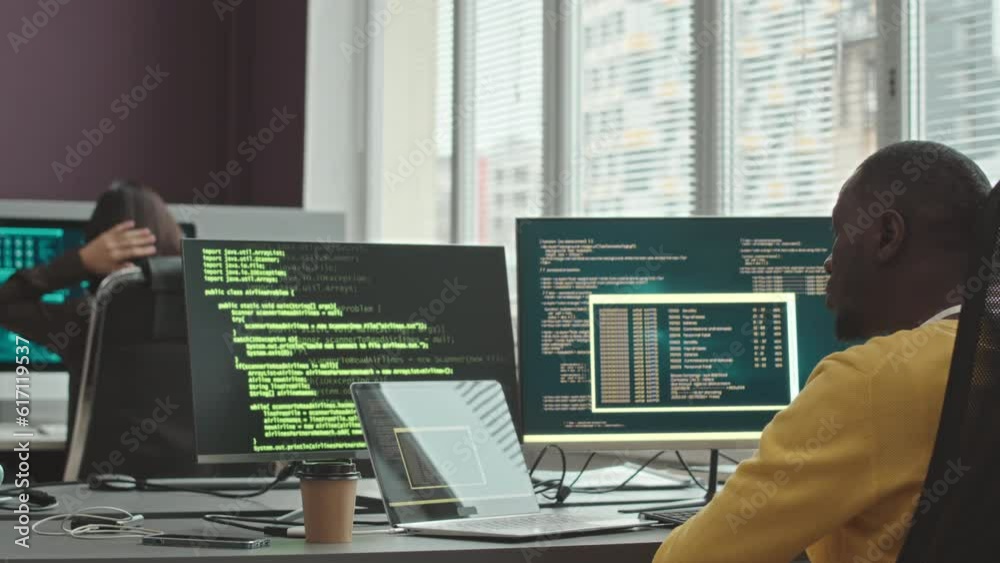 Back view of multiethnic couple of software developers program coding and monitoring information on cybersecurity servers sitting by computer monitors in office