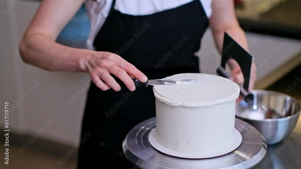 Concentrated blonde woman in black apron busy with spreading white mastic around round cake standing in modern kitchen confectioner enjoying making delicious sugar free pastry at home