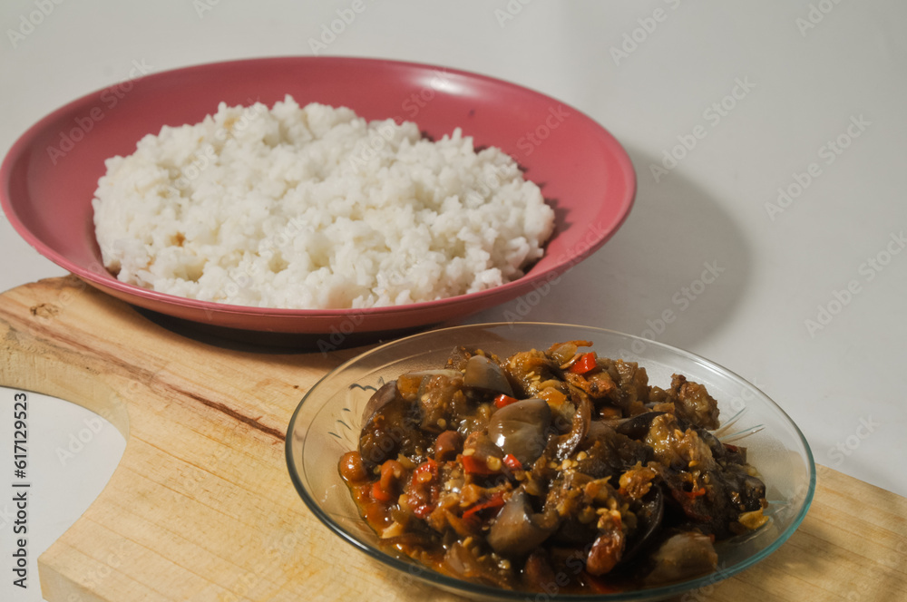 Stir-fried purple eggplant with anchovies and peanuts served in a small plate on a wooden cutting board and a plate of white rice isolated on a white background Stir-fried purple eggplant with anchovies and peanuts served in a small plate on a wooden cutting board and a plate of white rice isolated on a white background
