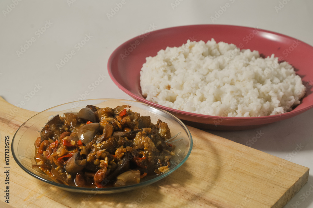 Stir-fried purple eggplant with anchovies and peanuts served in a small plate on a wooden cutting board and a plate of white rice isolated on a white background Stir-fried purple eggplant with anchovies and peanuts served in a small plate on a wooden cutting board and a plate of white rice isolated on a white background