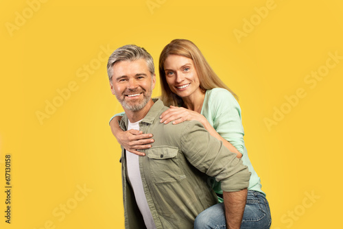 Excited european middle aged couple fooling together, happy woman piggybacking her husband, yellow background