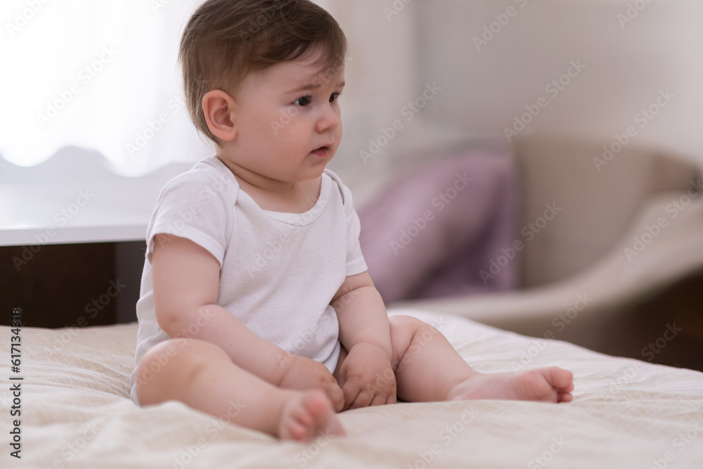 Cute chubby baby boy in white bodysuit sitting on soft bed looking to side adorable barefoot kid relaxing in bedroom at home after waking up in morning Cute chubby baby boy in white bodysuit sitting on soft bed looking to side adorable barefoot kid relaxing in bedroom at home after waking up in morning