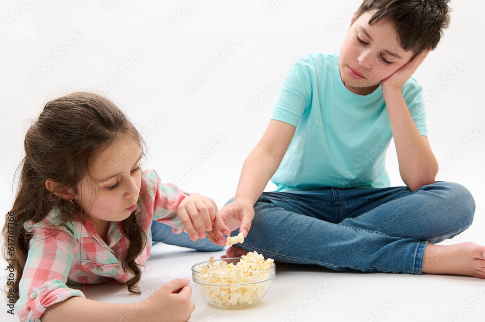 Caucasian happy primary school kids - teenager boy, brother and his younger sister, adorable litttle child girl eating popcorn, isolated on white studio background. Bonding. Family. Lifestyle Leisure Caucasian happy primary school kids - teenager boy, brother and his younger sister, adorable litttle child girl eating popcorn, isolated on white studio background. Bonding. Family. Lifestyle Leisure