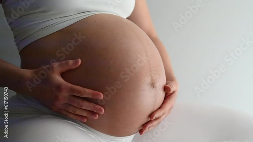 Midsection of heavily pregnant baby bump held by hands of woman bouncing on exercise ball. Side view. White background. Bright shot.