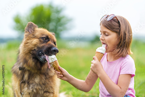 Cute little girl eating ice cream and feeding it to her shepherd dog sitting on the grass in the park in summer close up. High quality photo, blurred background.