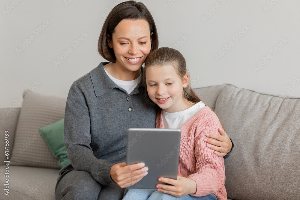 Glad millennial caucasian woman hugging teen daughter, have video call, read message on tablet on sofa
