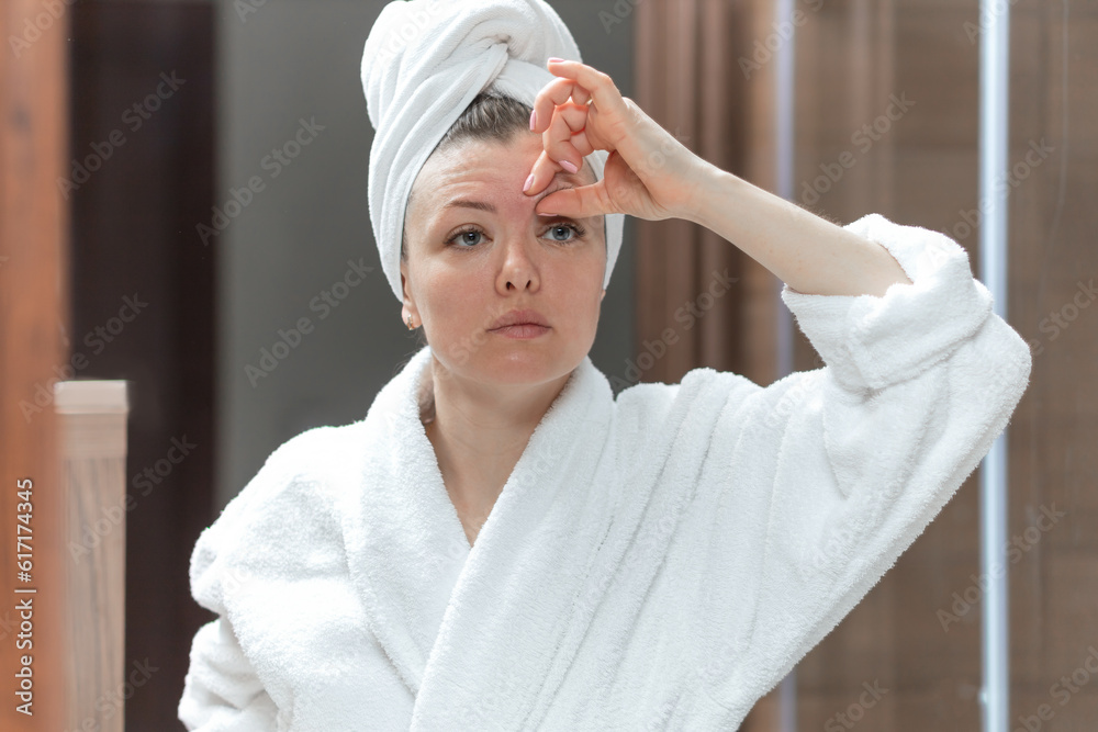 attractive young woman in white coat doing herself a facial lifting  massage  in mirror. Female take care for skin in bathroom, removing mimic wrinkles. personal self care. Wellness, Beauty concept