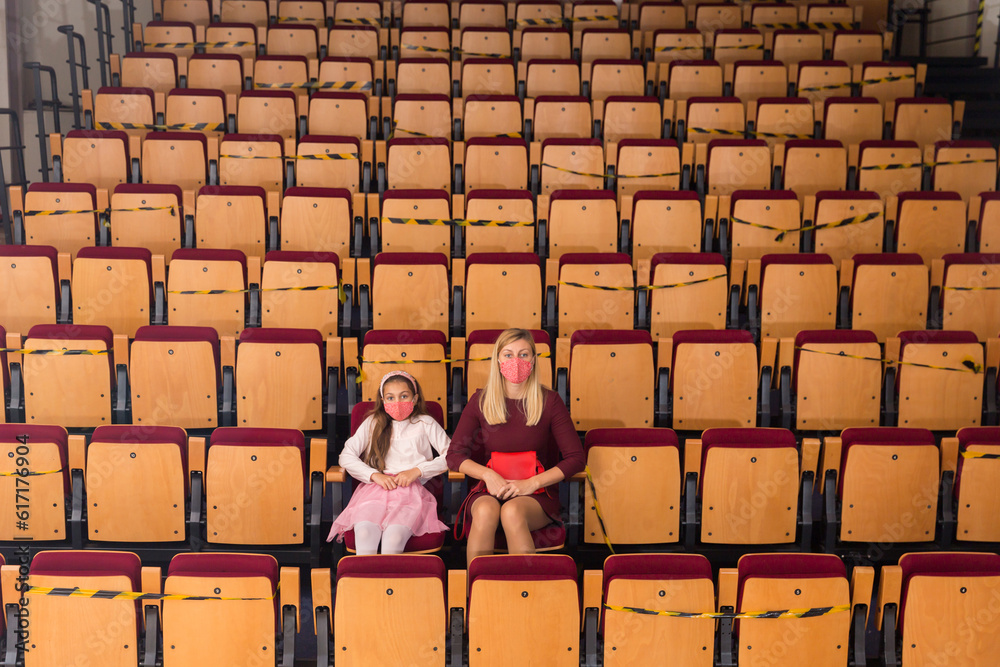 Φωτογραφία Young woman with preteen daughter wearing protective masks sitting in empty theater hall, watching play