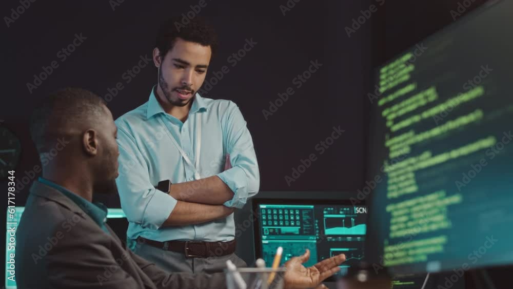 Medium shot of two diverse professional cybersecurity workers analyzing data on computer monitor and having discussion working together in agency at night