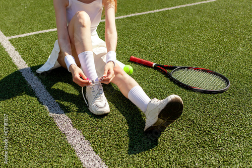 young girl tennis player in white sports uniform ties her shoelaces on sneakers on green court, woman coach