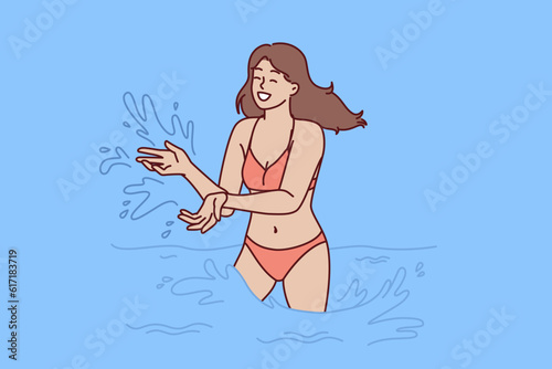 Woman in bikini swims in sea and splashes with water enjoying summer travel or trip to aquapark. Happy girl in swimsuit stands knee-deep in sea during vacation on tropical island with hot climate