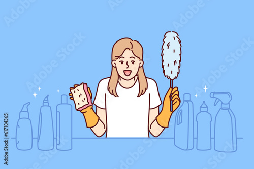 Cheerful woman housekeeper screams and holds cleaning brush in hands, standing near table with detergents. Girl housekeeper rejoices in good chemicals for cleaning in apartment or house