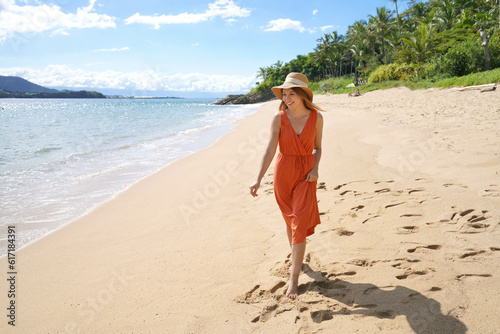 Young woman with orange sundress and straw hat walking on the sand beach photo